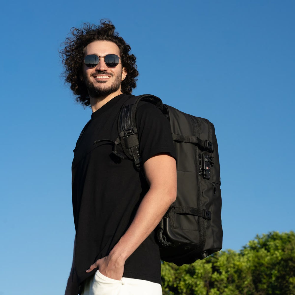 Homem usando a Backvac Pro Max preta como mochila sob um céu azul, com o cadeado TSA e uma rodinha à mostra